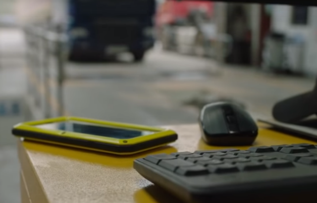 A mobile phone sitting on a desk next to a mouse and keyboard. A heavy vehicle testing pit in the background.